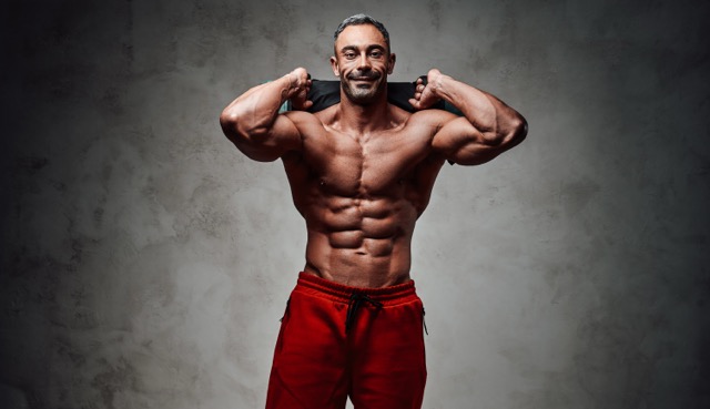 Buy Cardarine GW-501516 Tablets Online: Masculine sportsman isolated on the grey background wearing sport clothes while holding a sand bag up and smiling